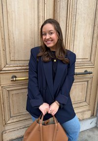 Femme souriante en blazer bleu marine et jeans tenant un grand sac en cuir marron, debout devant une porte en bois texturée.
