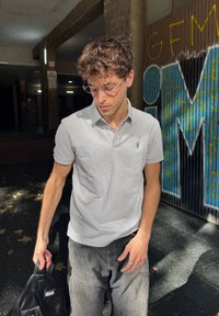 Polo shirt rayé gris et blanc, à manches courtes, avec un petit logo. Porté avec un jean gris clair, tenant un sac noir. Fond urbain avec des graffitis.