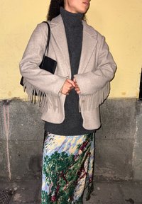 Άτομο που φοράει μπεζ μπουφάν με φουντάκια πάνω από σκούρο γκρι ζιβάγκο και πολύχρωμη φούστα με σχέδια, κρατώντας μαύρη τσάντα, στέκεται δίπλα σε τοίχο με υφή.