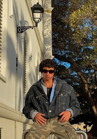 Chaqueta de mezclilla, azul oscuro, con bolsillos prominentes; combinada con una camisa de rayas. Pantalones de camuflaje, auriculares, gafas de sol, al aire libre bajo los árboles.