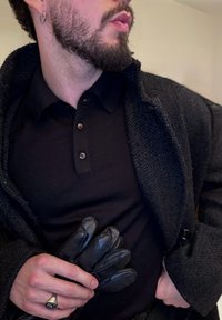 Personne barbu portant une chemise noire et une veste texturée foncée, tenant des gants en cuir noir dans une main, portant une bague et une boucle d'oreille en anneau.