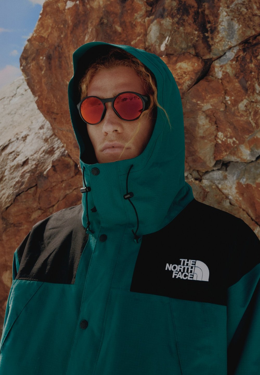 Τζάκετ σε τιρκουάζ απόχρωση με μαύρες λεπτομέρειες, ψηλό γιακά και ρυθμιζόμενα κορδόνια. Φέρει το λογότυπο "The North Face" στο στήθος. Το μοντέλο φοράει κόκκινα γυαλιά ηλίου.