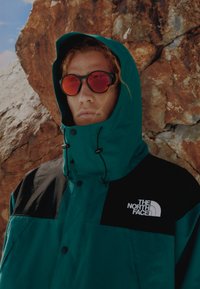 Τζάκετ σε τιρκουάζ απόχρωση με μαύρες λεπτομέρειες, ψηλό γιακά και ρυθμιζόμενα κορδόνια. Φέρει το λογότυπο "The North Face" στο στήθος. Το μοντέλο φοράει κόκκινα γυαλιά ηλίου.