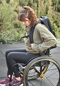 Schwarzer Rollstuhl mit goldenen Akzenten, Benutzer in einer grünen Fleecejacke und schwarzen Leggings, der einen schwarzen Rucksack anpasst, im Freien.