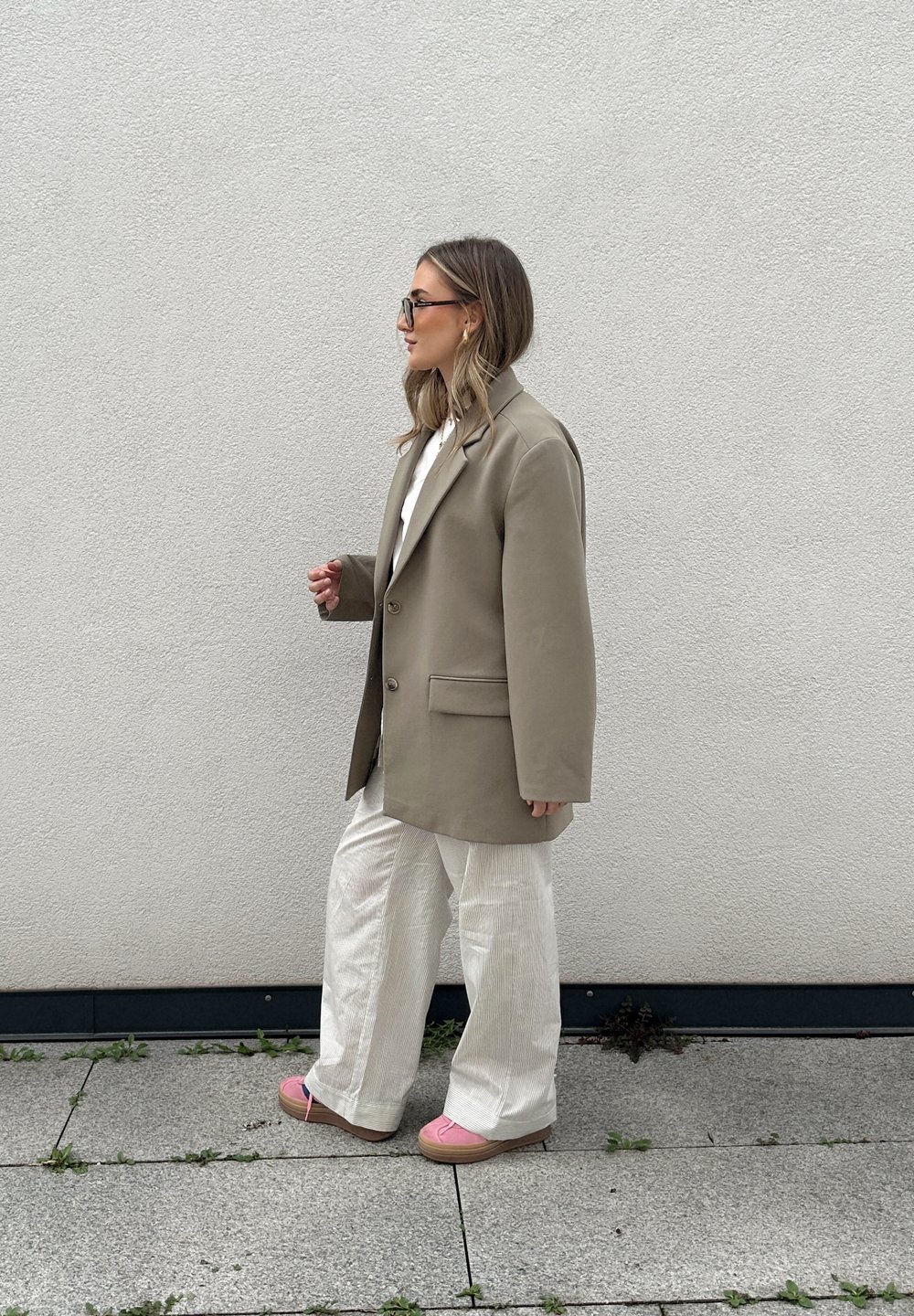 Blazer beige oversize con un bottone, abbinato a pantaloni bianchi a pieghe e scarpe con plateau rosa, contro uno sfondo di muro uniforme.
