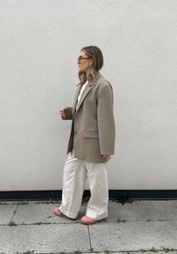 Blazer beige oversize con un bottone, abbinato a pantaloni bianchi a pieghe e scarpe con plateau rosa, contro uno sfondo di muro uniforme.