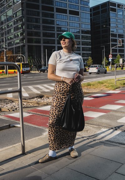 Falda maxi de estampado de leopardo, blusa blanca de manga corta, gorra verde, bolso negro, zapatos marrones tipo slip-on con calcetines blancos. Escenario urbano con edificios.