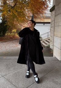 Černý texturovaný trenčkot přes černé šaty s třásněmi, doplněné černými legínami a robustními černými botami s bílými prvky. Na pozadí podzimní listí.