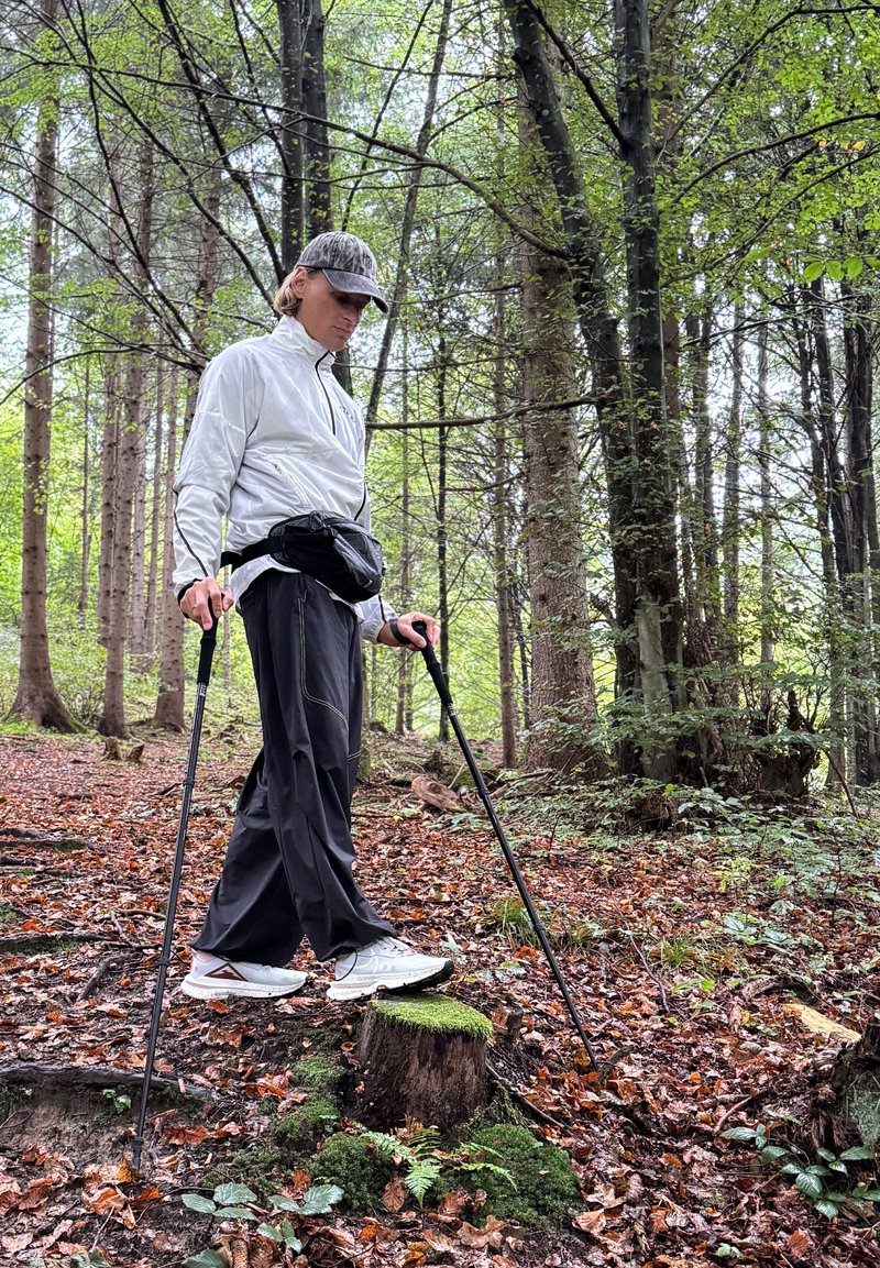 Vaeltaja valkoisessa takissa ja mustissa housuissa käyttää vaellussauvoja metsässä. Tausta sisältää puita, kasvillisuutta ja pudonneita lehtiä.