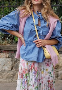 Bluse mit blau-weißen Streifen und Bindedetail, kombiniert mit einer rosa Strickjacke und einem plissierten Blumenkleid. Gelbe Umhängetasche.