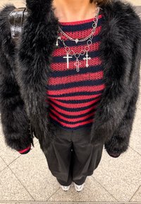 Veste en fausse fourrure noire superposée sur un haut en tricot rayé rouge et marine avec des colliers croix argentés. Un pantalon noir ample complète la tenue.