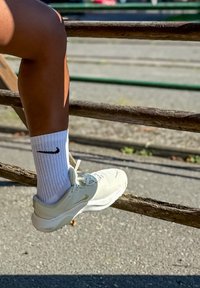 Zapatilla deportiva Nike en color crema claro con parte superior de malla texturizada y un acento dorado, combinada con calcetines blancos de canalé que muestran un logo negro de Nike.