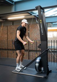 Concept2 sõudmismasin musta värvi hõbedaste detailidega; isik kasutab reguleeritavaid takistusega kaableid, kannab musti lühikesi pükse ja nokamütsi.