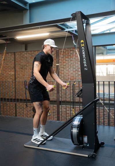 Máquina de remo Concept2 en negro con detalles plateados; persona utilizando cables de resistencia ajustable, vistiendo pantalones cortos negros y gorra.