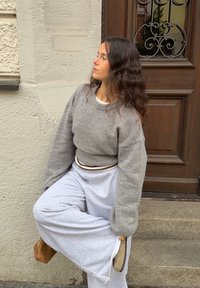 Grå, oversized sweater med brede ærmer; kombineret med løstsiddende lysegrå joggingbukser og brunt fodtøj. Baggrunden viser en trædør.