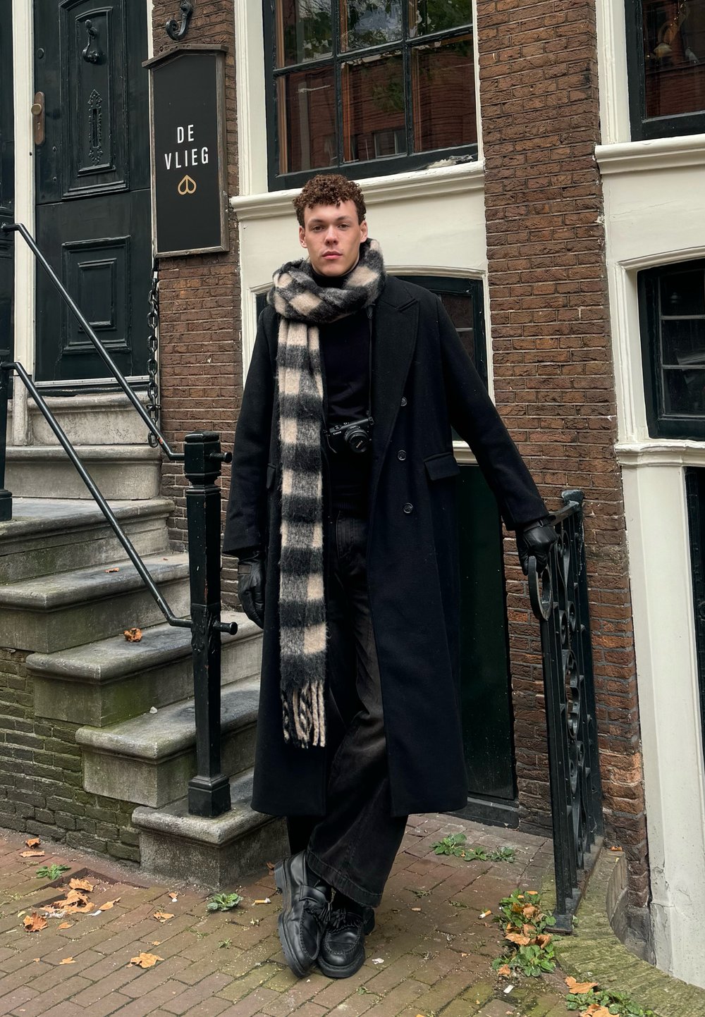 Μαύρο μακρύ παλτό πάνω από μαύρο πουλόβερ, patterned κρεμάστρα σε μαύρο και κρέμα, μαύρα τζιν και μαύρα παπούτσια. Στηρίζει σε τοίχο από το ύβλα, σκάλες και κάγκελο.