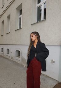 Schwarzer übergroßer Blazer, eng anliegendes schwarzes Top und hoch taillierte burgunderrote Schlaghosen. Glatte Texturen, lockere urbane Umgebung mit grauen Wänden.