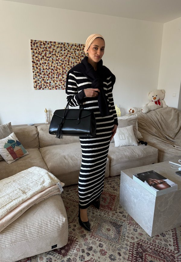 Mujer con vestido de rayas negras y blancas, pañuelo beige en la cabeza y tacones negros sosteniendo un bolso negro en un salón.