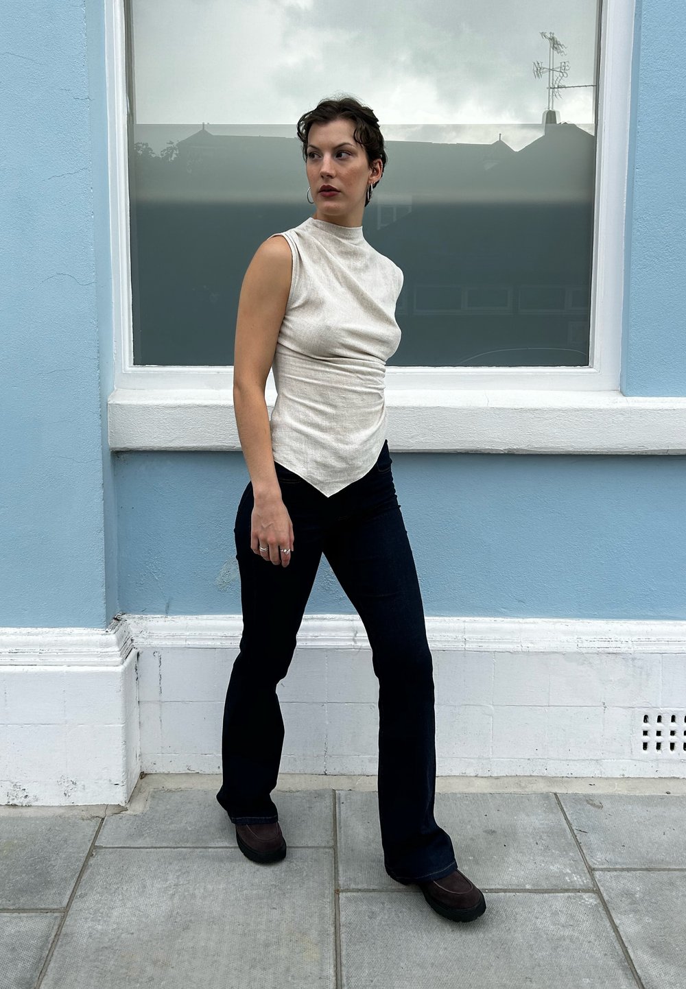 Αμάνικη μπεζ μπλούζα με υφασμάτινη υφή και ασύμμετρο στρίφωμα, συνδυασμένη με σκούρα καμπάνα τζιν και καφέ παπούτσια, με φόντο έναν μπλε τοίχο.