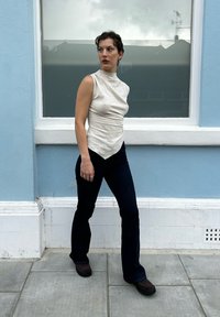 Αμάνικη μπεζ μπλούζα με υφασμάτινη υφή και ασύμμετρο στρίφωμα, συνδυασμένη με σκούρα καμπάνα τζιν και καφέ παπούτσια, με φόντο έναν μπλε τοίχο.