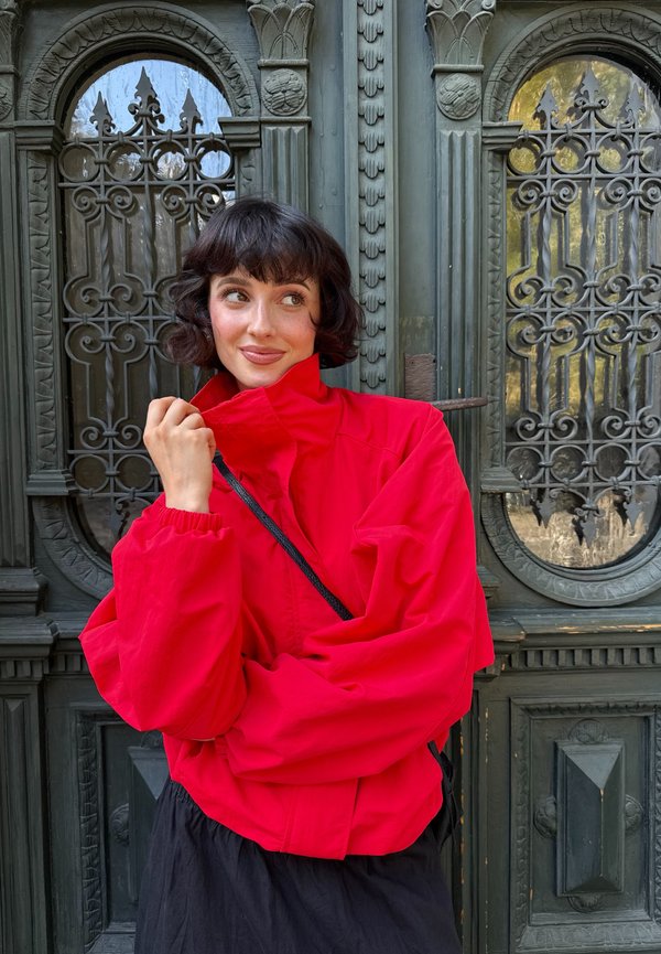 Joven mujer con cabello corto y oscuro que lleva una chaqueta roja brillante, está frente a unas puertas de metal verde oscuro ornamentadas, mirando hacia un lado y sonriendo.