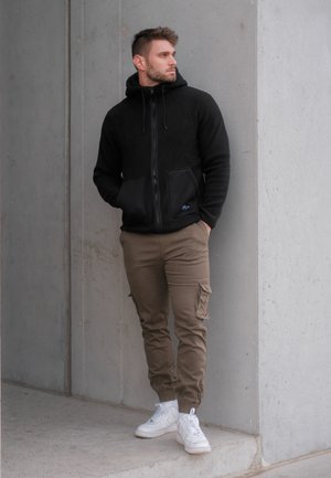 Sudadera con cremallera de forro polar negra con dos bolsillos frontales, combinada con pantalones cargo beige y zapatillas blancas, frente a una pared de hormigón.