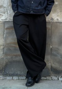 Vide sorte bukser i glat stof, kombineret med sorte sko. Modellen læner sig op ad en tekstureret stenvæg.