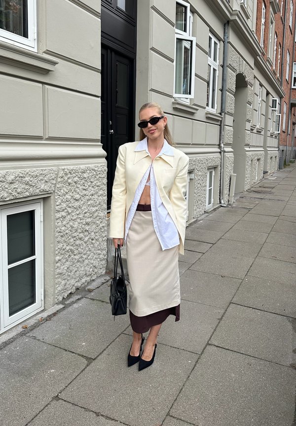 Una mujer con gafas de sol, americana y falda de color crema, sostiene un bolso negro en una acera de la ciudad, junto a una hilera de edificios.