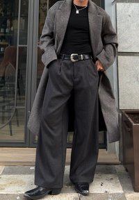 Mann iført lang grå frakk, svart skjorte, vide mørkegrå bukser, svart skinnbelte med dekorativ spenne, og svarte sko utenfor en bygning.