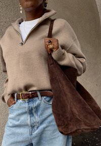 Άτομο που φοράει μπεζ πουλόβερ με φερμουάρ στο λαιμό, ανοιχτό μπλε τζιν, καφέ δερμάτινη ζώνη, κ ρατάει μεγάλη καφέ σουέντ τσάντα tote και φορά χρυσά δαχτυλίδια και σκουλαρίκια.