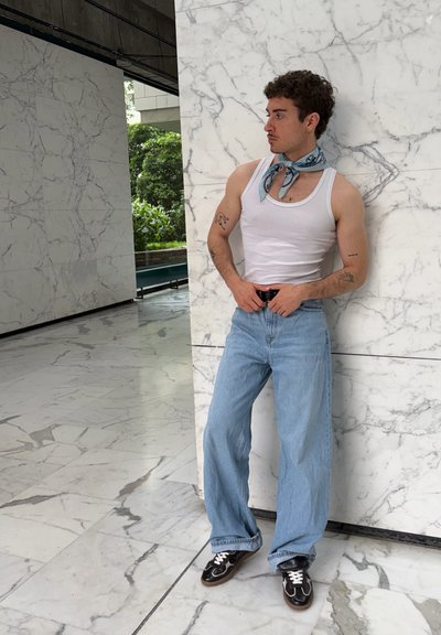 Hombre con pelo rizado está de pie frente a una pared de mármol blanco, lleva una camiseta de tirantes blanca, vaqueros azul claro, zapatillas negras y una bufanda azul en el cuello.