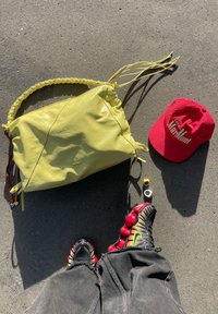 Άτομο που φοράει μαύρα και κόκκινα αθλητικά παπούτσια Nike και μαύρο τζιν, στέκεται κοντά σε μια κίτρινη τσάντα, κόκκινο καπέλο Maramont και δύο μικρές ηλεκτρονικές συσκευές πάνω σε άσφαλτο.