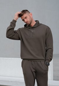 Sweat à capuche vert olive foncé avec une poche kangourou et des cordons de serrage, assorti à un pantalon de jogging. Présente un logo sur la poitrine et une montre au poignet.