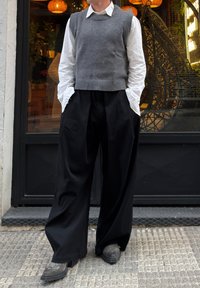 Pull sans manches gris en maille porté sur une chemise blanche, associé à un pantalon large noir et des chaussures grises à motif texturé.