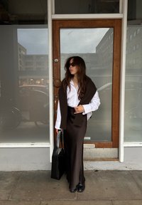 Pulover tricotat maro așezat peste o cămașă albă cu nasturi, asortat cu o fustă satinată maro. O geantă neagră și pantofi cu platformă completează ținuta.