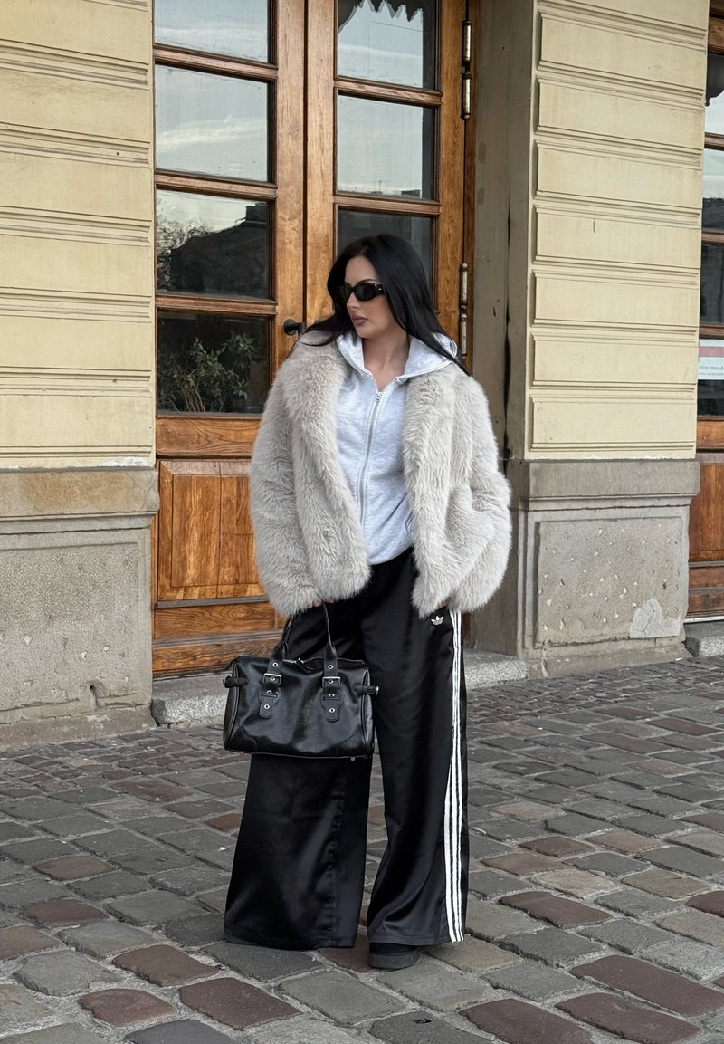 Chaqueta de pelo sintético gris, sudadera con cremallera gris, pantalones anchos de satén negros con rayas blancas y un bolso negro. De pie cerca de puertas de madera.