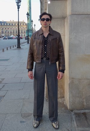 Chaqueta de cuero marrón, camisa negra con lunares, pantalones a rayas grises, zapatos con estampado de serpiente, gafas de sol; de pie contra un muro de piedra.
