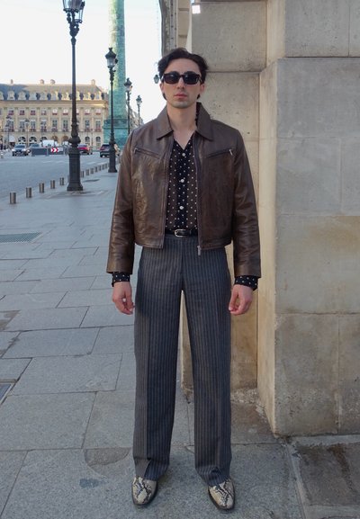 Chaqueta de cuero marrón, camisa negra con lunares, pantalones a rayas grises, zapatos con estampado de serpiente, gafas de sol; de pie contra un muro de piedra.