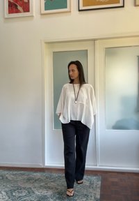 Biely ľahký ľanový top s voľným strihom, spárovaný s čiernymi pohodlnými nohavicami. Stojí na vzorovanom koberci v minimalistickom interiéri.