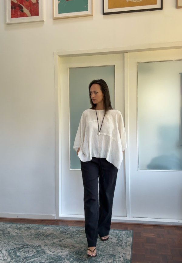 Leggero top in lino bianco con vestibilità ampia, abbinato a pantaloni neri rilassati. In piedi su un tappeto a motivi in un interno minimalista.