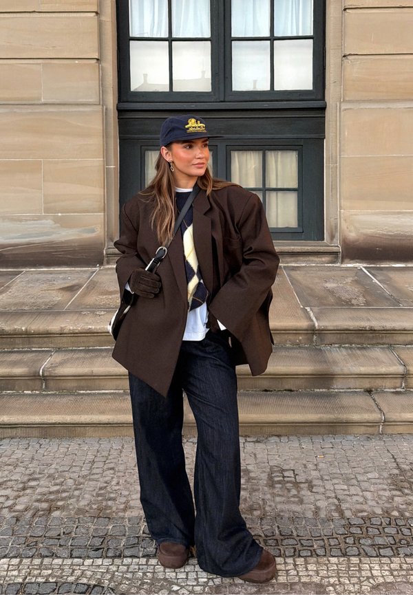 Femeie purtând un sacou larg de culoare maro închis, vestă pulover cu model argyle, șapcă bleumarin, pantaloni largi și mănuși, stând pe trepte de piatră în afara unei clădiri.