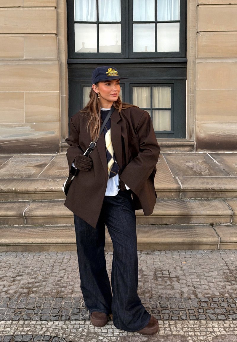 Femme portant un blazer oversize marron foncé, un gilet en maille à motif losange, une casquette marine, un pantalon large et des gants, debout sur des marches en pierre devant un bâtiment.