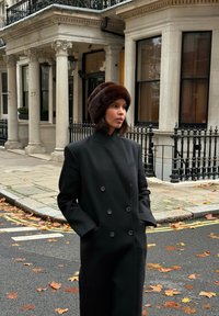 Fekete gyapjú ballonkabát dupla soros gombolással, széles hajtókákkal és oldalzsebekkel. Sötét szőrmekalappal párosítva, utcai háttér előtt.