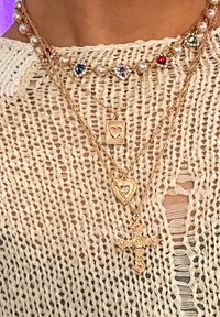 Στρωματωμένα χρυσά κολιέ που περιλαμβάνουν μαργαρίτες και πολύχρωμους πολύτιμους λίθους, συμπεριλαμβανομένου ενός κρεμαστού κόκκινου καρδιού και ενός διακοσμητικού σταυρού, σε φόντο από πλεκτό τοπ.