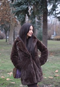 Mladá žena v hnedom huňatom kabáte stojí v parku s rukami v kapsách na pozadí zelenej trávy a vysokých stromov na jeseň.