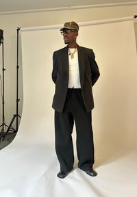 Mann, der eine Camouflage-Cap, Brille, goldene Halsketten, ein weißes Hemd, einen schwarzen Blazer, weite schwarze Hosen und Zehenstegschuhe trägt, steht in einem Studio mit weißem Hintergrund.