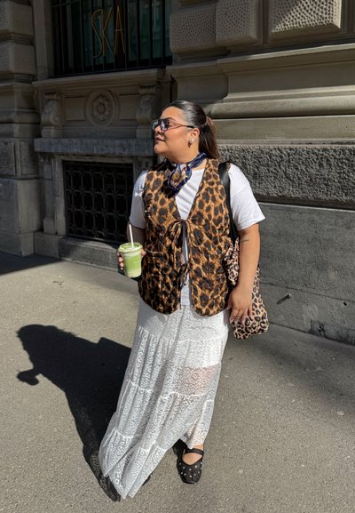 Chaleco de estampado de leopardo marrón sobre una camiseta blanca, combinado con una falda blanca transparente. Accesorios incluyen una bufanda con estampado y un bolso de estampado de leopardo.