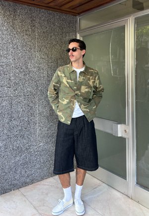 Hombre con gafas de sol, chaqueta de camuflaje, camisa blanca, pantalones cortos largos negros, calcetines blancos y zapatillas blancas, está de pie en el interior junto a una pared gris y una puerta de cristal.