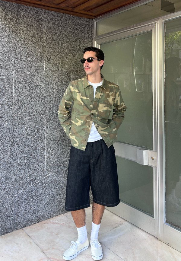 Homme portant des lunettes de soleil, une veste camouflage, une chemise blanche, un short long noir, des chaussettes blanches et des baskets blanches, debout à l'intérieur près d'un mur gris et d'une porte en verre.