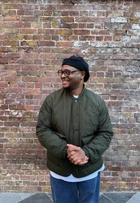 Veste matelassée verte avec un col rond, béret noir et lunettes à monture transparente, associée à une chemise blanche et un jean bleu. Fond en briques texturées.